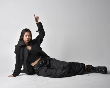 Full length portrait of pretty young asian girl wearing black tank top, utilitarian  pants, leather boots and large dark coat. Sitting pose with gestural hand movements,  isolated on  studio background.
