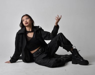 Full length portrait of pretty young asian girl wearing black tank top, utilitarian  pants, leather boots and large dark coat. Sitting pose with gestural hand movements,  isolated on  studio background.