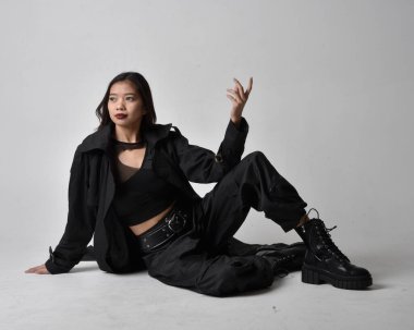 Full length portrait of pretty young asian girl wearing black tank top, utilitarian  pants, leather boots and large dark coat. Sitting pose with gestural hand movements,  isolated on  studio background.