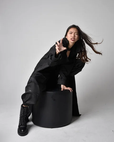 全长肖像漂亮年轻的亚洲女孩穿着黑色的坦克顶部 实用的裤子 皮靴和大的深色外套 手部有手势动作的坐姿 独立于工作室背景 — 图库照片