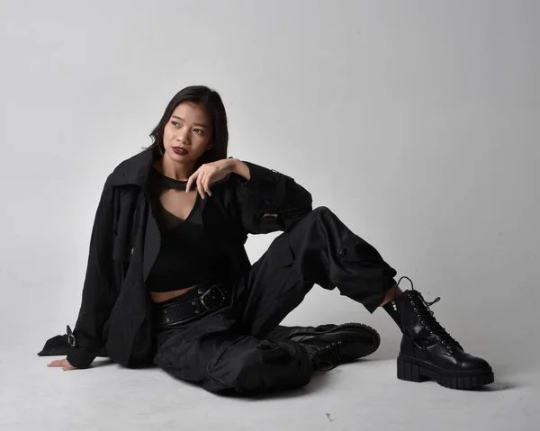 全长肖像漂亮年轻的亚洲女孩穿着黑色的坦克顶部 实用的裤子 皮靴和大的深色外套 手部有手势动作的坐姿 独立于工作室背景 — 图库照片