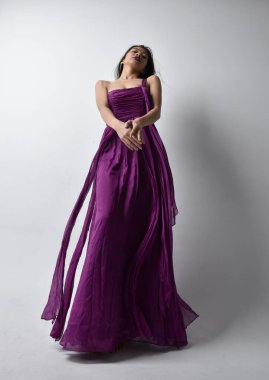 Full length  portrait of pretty brunette asian girl wearing purple flowing  gown. Standing pose in low angle on on studio background.