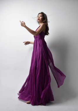 Full length  portrait of pretty brunette asian girl wearing purple flowing  gown. Standing pose in low angle on on studio background.