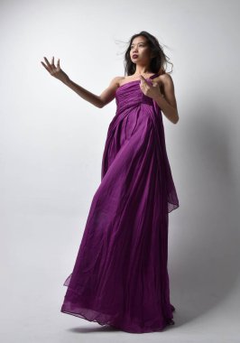 Full length  portrait of pretty brunette asian girl wearing purple flowing  gown. Standing pose in low angle on on studio background.