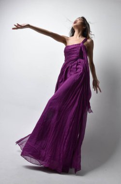 Full length  portrait of pretty brunette asian girl wearing purple flowing  gown. Standing pose in low angle on on studio background.