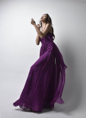 Full length  portrait of pretty brunette asian girl wearing purple flowing  gown. Standing pose in low angle on on studio background.