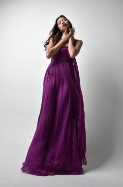 Full length  portrait of pretty brunette asian girl wearing purple flowing  gown. Standing pose in low angle on on studio background.
