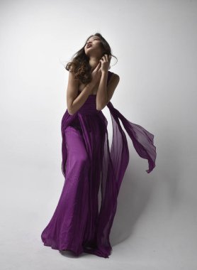 Full length  portrait of pretty brunette asian girl wearing purple flowing  gown. Standing pose in low angle on on studio background.