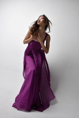 Full length  portrait of pretty brunette asian girl wearing purple flowing  gown. Standing pose in low angle on on studio background.