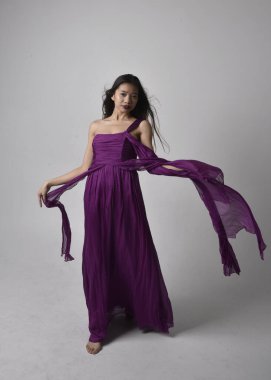 Full length  portrait of pretty brunette asian girl wearing purple flowing  gown. Standing, dancing pose on on studio background.