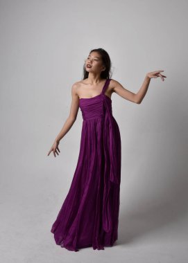 Full length  portrait of pretty brunette asian girl wearing purple flowing  gown. Standing, dancing pose on on studio background.