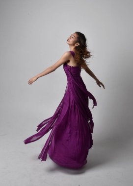 Full length  portrait of pretty brunette asian girl wearing purple flowing  gown. Standing, dancing pose on on studio background.