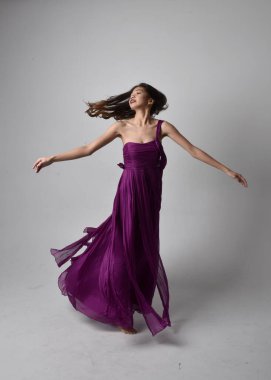 Full length  portrait of pretty brunette asian girl wearing purple flowing  gown. Standing, dancing pose on on studio background.