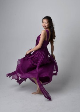 Full length  portrait of pretty brunette asian girl wearing purple flowing  gown. Standing, dancing pose on on studio background.