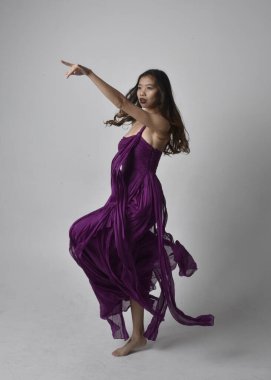 Full length  portrait of pretty brunette asian girl wearing purple flowing  gown. Standing, dancing pose on on studio background.