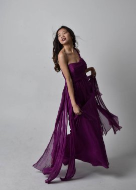 Full length  portrait of pretty brunette asian girl wearing purple flowing  gown. Standing, dancing pose on on studio background.