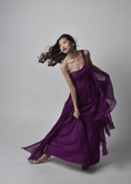 Full length  portrait of pretty brunette asian girl wearing purple flowing  gown. Standing, dancing pose on on studio background.