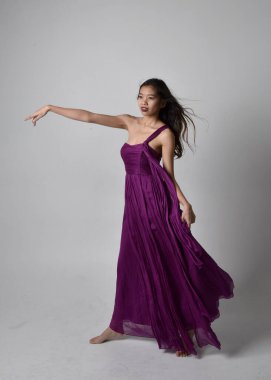 Full length  portrait of pretty brunette asian girl wearing purple flowing  gown. Standing, dancing pose on on studio background.