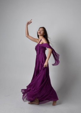 Full length  portrait of pretty brunette asian girl wearing purple flowing  gown. Standing, dancing pose on on studio background.