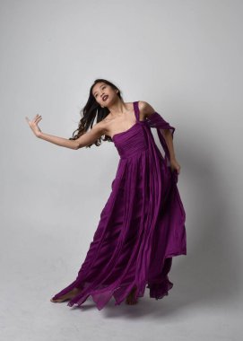 Full length  portrait of pretty brunette asian girl wearing purple flowing  gown. Standing, dancing pose on on studio background.