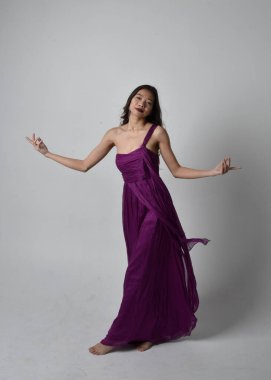 Full length  portrait of pretty brunette asian girl wearing purple flowing  gown. Standing, dancing pose on on studio background.
