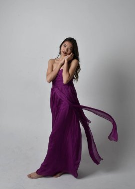 Full length  portrait of pretty brunette asian girl wearing purple flowing  gown. Standing, dancing pose on on studio background.