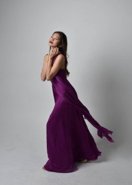 Full length  portrait of pretty brunette asian girl wearing purple flowing  gown. Standing, dancing pose on on studio background.