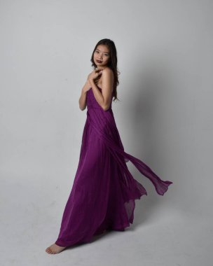 Full length  portrait of pretty brunette asian girl wearing purple flowing  gown. Standing, dancing pose on on studio background.