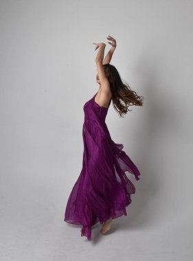 Full length  portrait of pretty brunette asian girl wearing purple flowing  gown. Standing, dancing pose on on studio background.