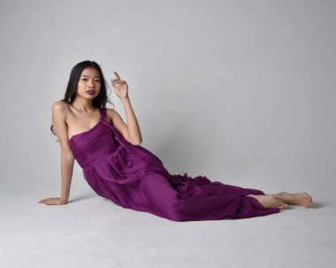 Full length  portrait of pretty brunette asian girl wearing purple flowing  gown. Sitting pose on on studio background.