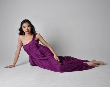 Full length  portrait of pretty brunette asian girl wearing purple flowing  gown. Sitting pose on on studio background.