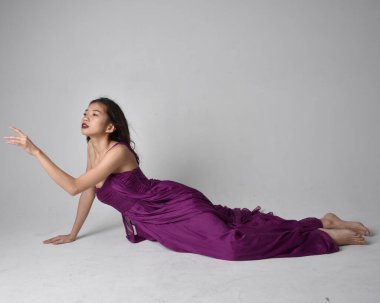 Full length  portrait of pretty brunette asian girl wearing purple flowing  gown. Sitting pose on on studio background.
