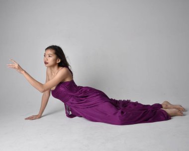Full length  portrait of pretty brunette asian girl wearing purple flowing  gown. Sitting pose on on studio background.