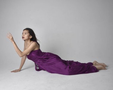 Full length  portrait of pretty brunette asian girl wearing purple flowing  gown. Sitting pose on on studio background.