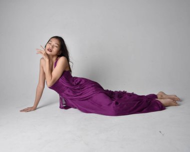 Full length  portrait of pretty brunette asian girl wearing purple flowing  gown. Sitting pose on on studio background.