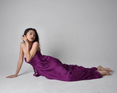 Full length  portrait of pretty brunette asian girl wearing purple flowing  gown. Sitting pose on on studio background.