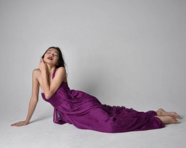Full length  portrait of pretty brunette asian girl wearing purple flowing  gown. Sitting pose on on studio background.