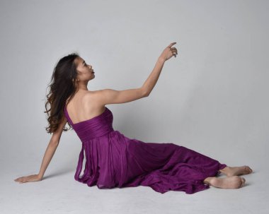 Full length  portrait of pretty brunette asian girl wearing purple flowing  gown. Sitting pose on on studio background.