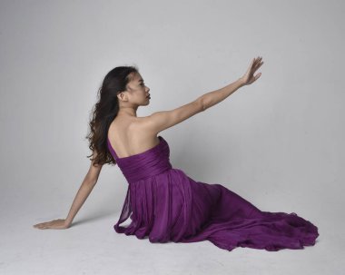 Full length  portrait of pretty brunette asian girl wearing purple flowing  gown. Sitting pose on on studio background.