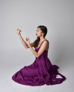 Full length  portrait of pretty brunette asian girl wearing purple flowing  gown. Sitting pose on on studio background.