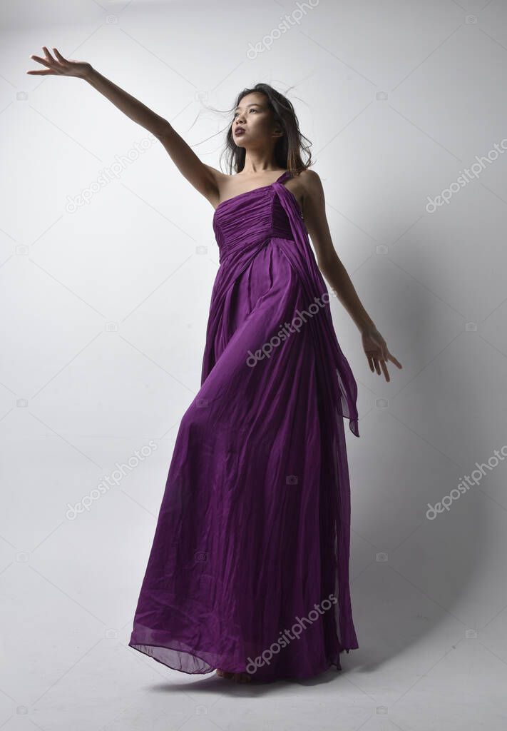 Full length  portrait of pretty brunette asian girl wearing purple flowing  gown. Standing pose in low angle on on studio background.