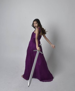 Full length  portrait of pretty brunette asian girl wearing purple flowing  gown. Standing pose holding a sword  on on studio background.