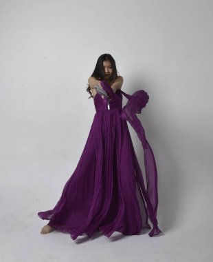 Full length  portrait of pretty brunette asian girl wearing purple flowing  gown. Standing pose holding a sword  on on studio background.