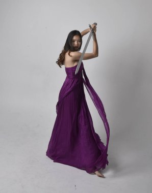 Full length  portrait of pretty brunette asian girl wearing purple flowing  gown. Standing pose holding a sword  on on studio background.