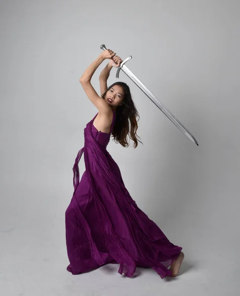 Full length  portrait of pretty brunette asian girl wearing purple flowing  gown. Standing pose holding a sword  on on studio background.