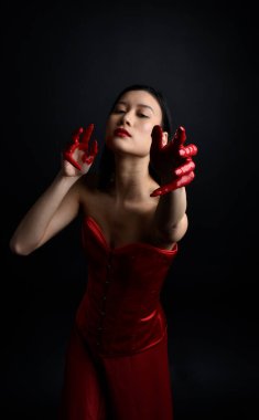 Kırmızı korse giyen güzel Asyalı bir kadının yakın plan portresi, kanlı boyayla kaplanmış, Gotik korku tarzında, yaratıcı el hareketleriyle koyu siyah stüdyo arka planına izole edilmiş..