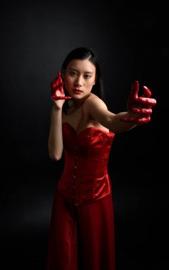 Kırmızı korse giyen güzel Asyalı bir kadının yakın plan portresi, kanlı boyayla kaplanmış, Gotik korku tarzında, yaratıcı el hareketleriyle koyu siyah stüdyo arka planına izole edilmiş..