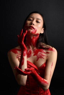 Kırmızı korse giyen güzel Asyalı bir kadının yakın plan portresi, kanlı boyayla kaplanmış, Gotik korku tarzında, yaratıcı el hareketleriyle koyu siyah stüdyo arka planına izole edilmiş..