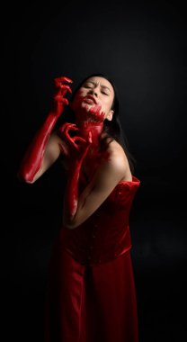 Kırmızı korse giyen güzel Asyalı bir kadının yakın plan portresi, kanlı boyayla kaplanmış, Gotik korku tarzında, yaratıcı el hareketleriyle koyu siyah stüdyo arka planına izole edilmiş..