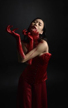 Kırmızı korse giyen güzel Asyalı bir kadının yakın plan portresi, kanlı boyayla kaplanmış, Gotik korku tarzında, yaratıcı el hareketleriyle koyu siyah stüdyo arka planına izole edilmiş..